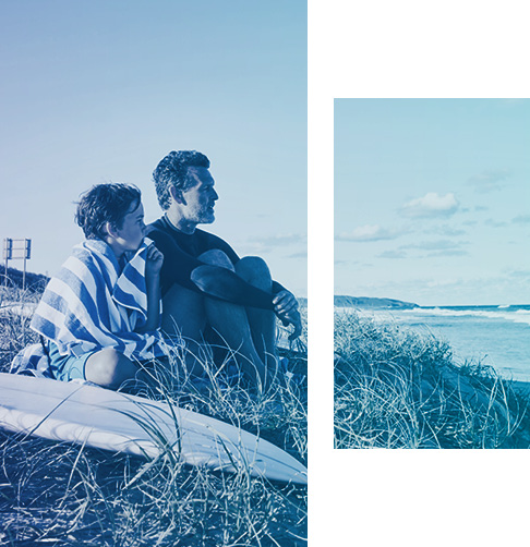 Un garçon et un homme assis sur la plage, à côté d’une planche de surf, regardant l’océan 